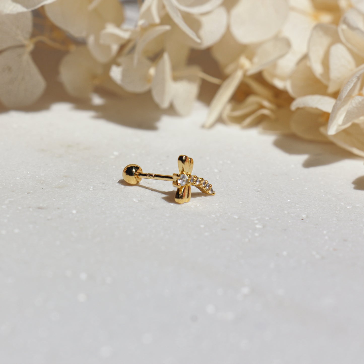 Gold dragonfly earring with a cubic zirconia on a white surface with beige flowers in the background