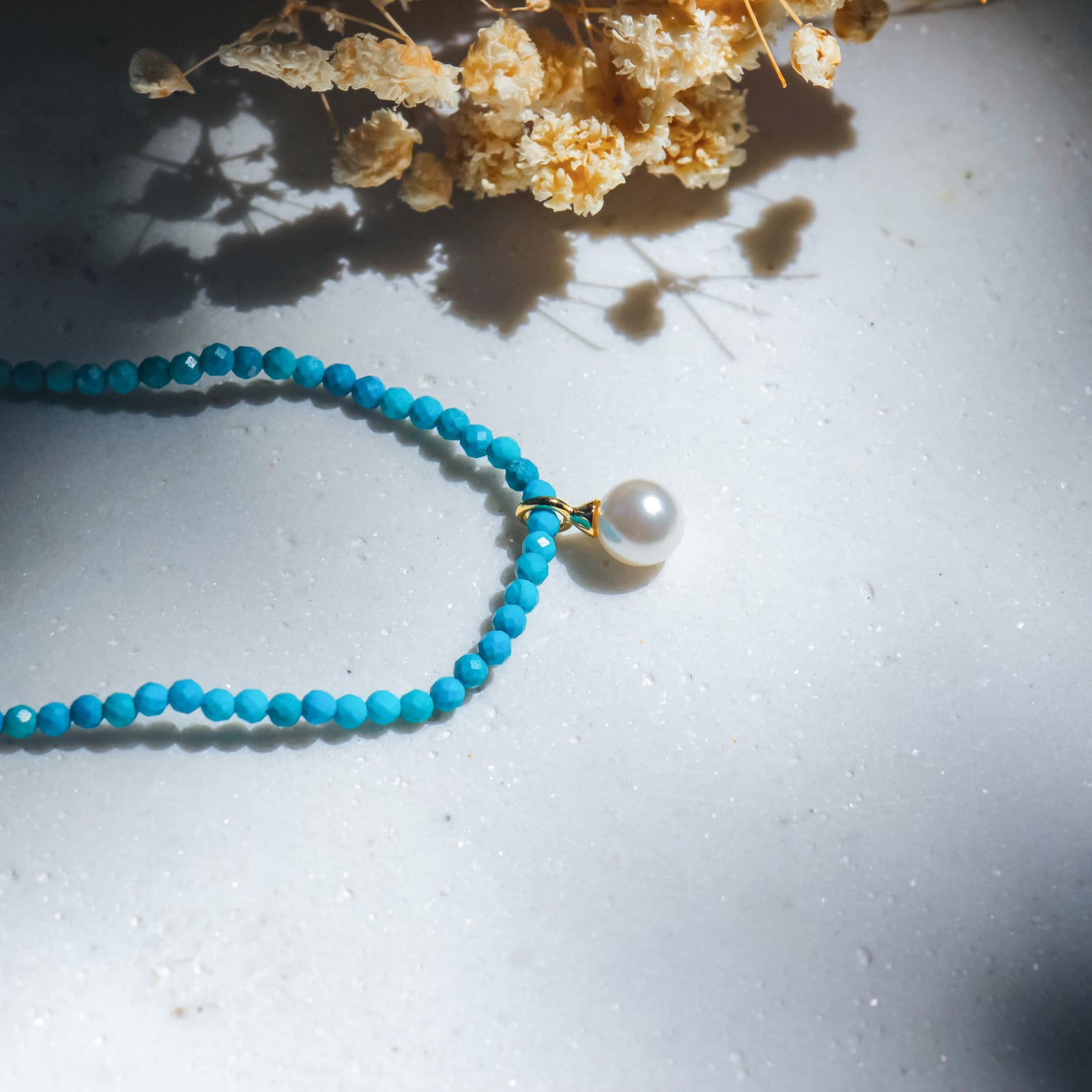 Turquoise beaded necklace with a pearl pendant on a light surface with dried flowers.