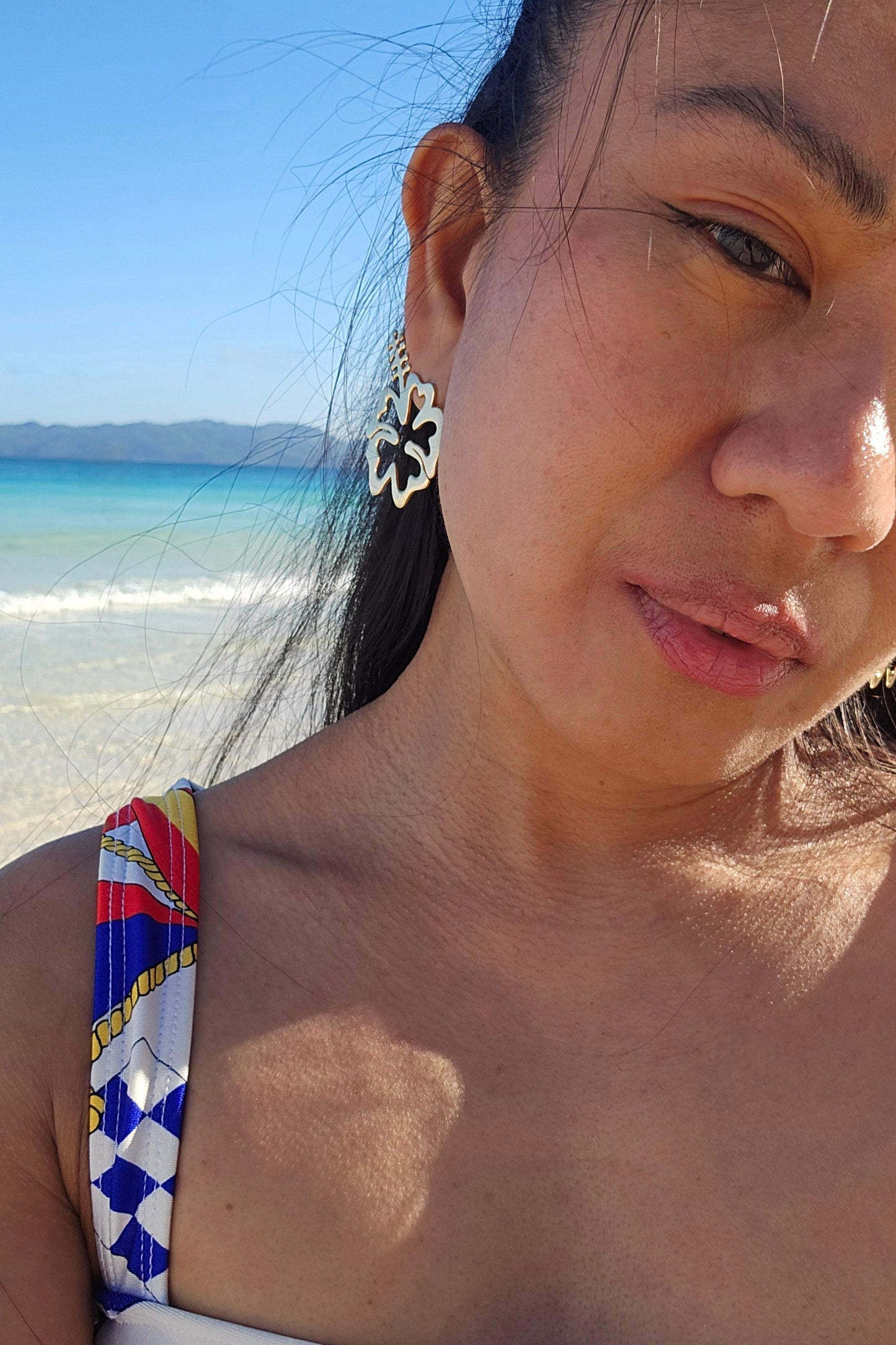 Woman wearing a colorful swimwear top with a beach in the background and big hibiscus earrings in 18K gold dipped Silver 925