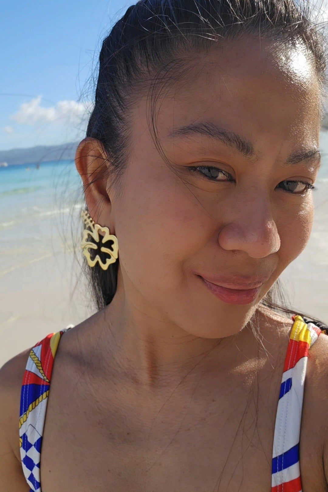 Woman wearing a colorful swimwear top with a beach in the background and big hibiscus earrings in 18K gold dipped Silver 925