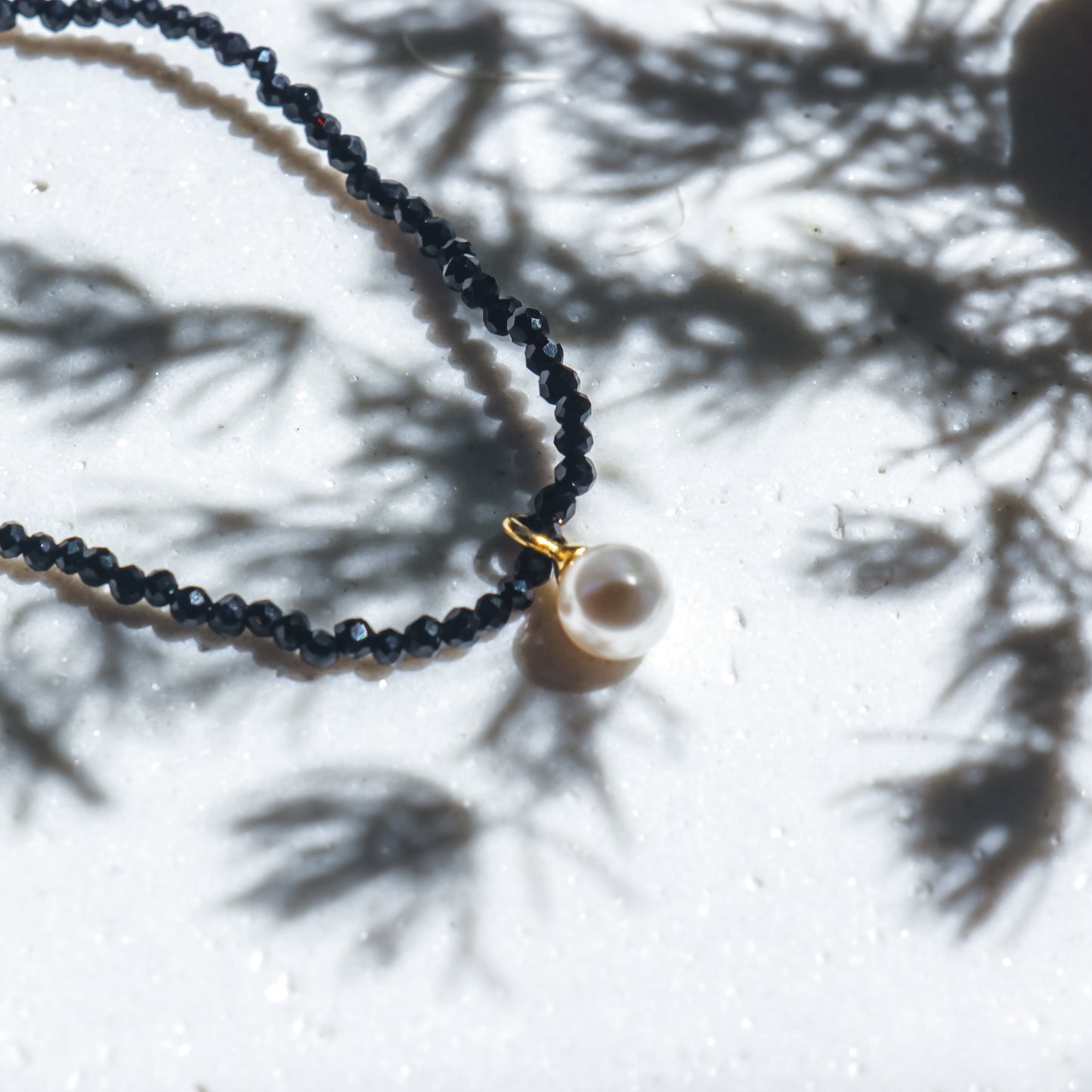 Black beaded necklace with a gold clasp and pearl pendant on a white surface with shadows.